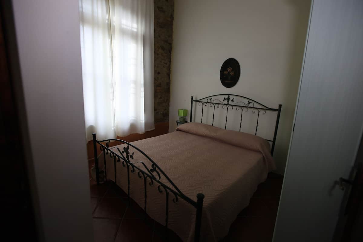 Een slaapkamer met een metalen bedframe, een beige sprei, een nachtkastje met een groen voorwerp, en een aan de muur hangend schilderij.