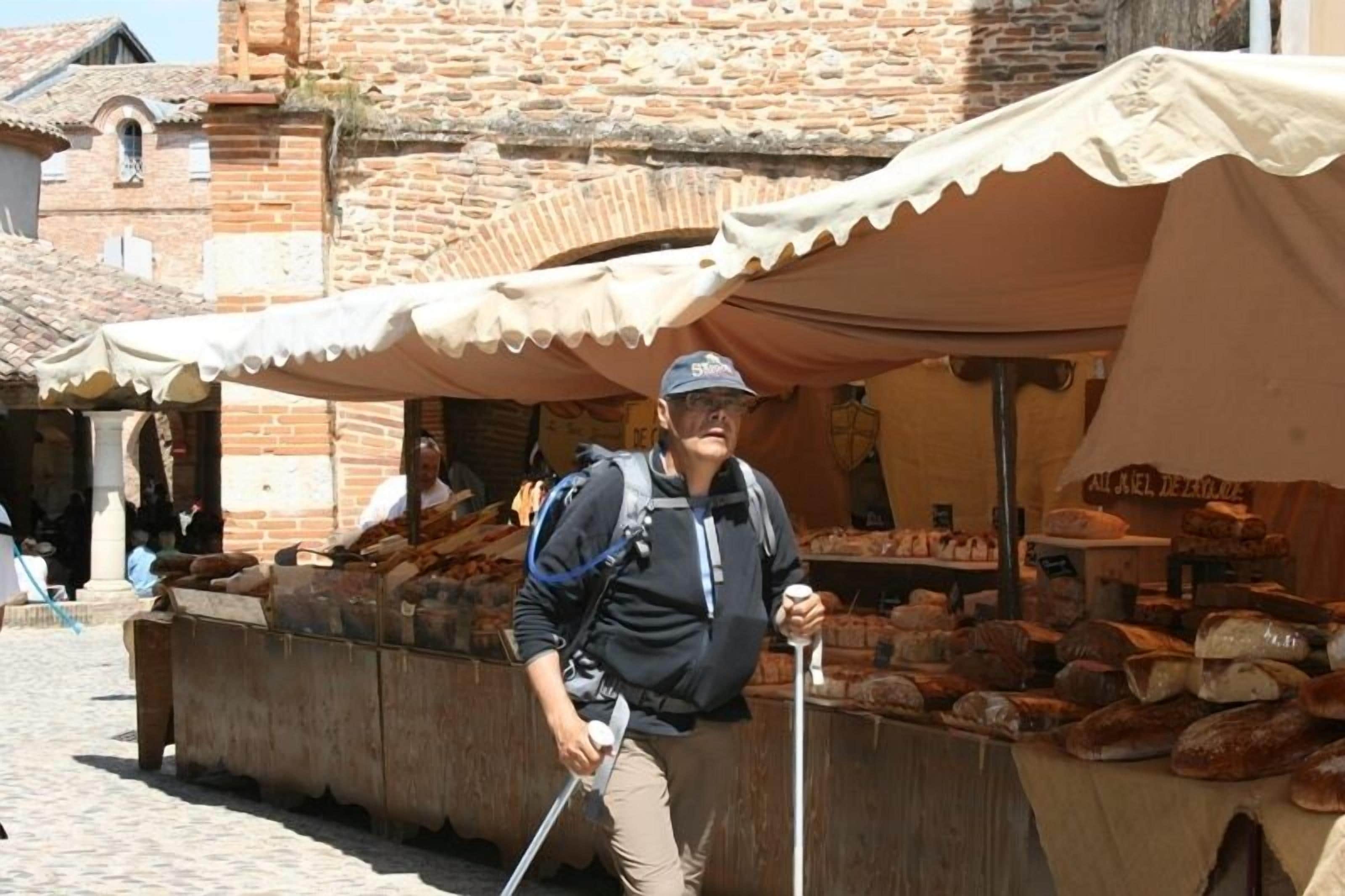 Une personne se tient devant un étal à pain, un sac à dos sur le dos et des bâtons de marche à la main, devant un bâtiment en pierre surmonté d’un toit en tuiles.