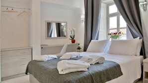A hotel room with a bed, a bedside table, a mirror, a window with curtains, a wardrobe, and a hanging rack with clothes. - Douskos Port House (Hydra)