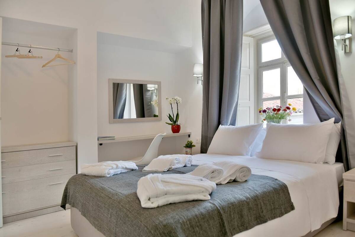 Quarto de hotel com cama, mesa de cabeceira, espelho, janela com cortinas, guarda-roupa e cabideiro com roupas.