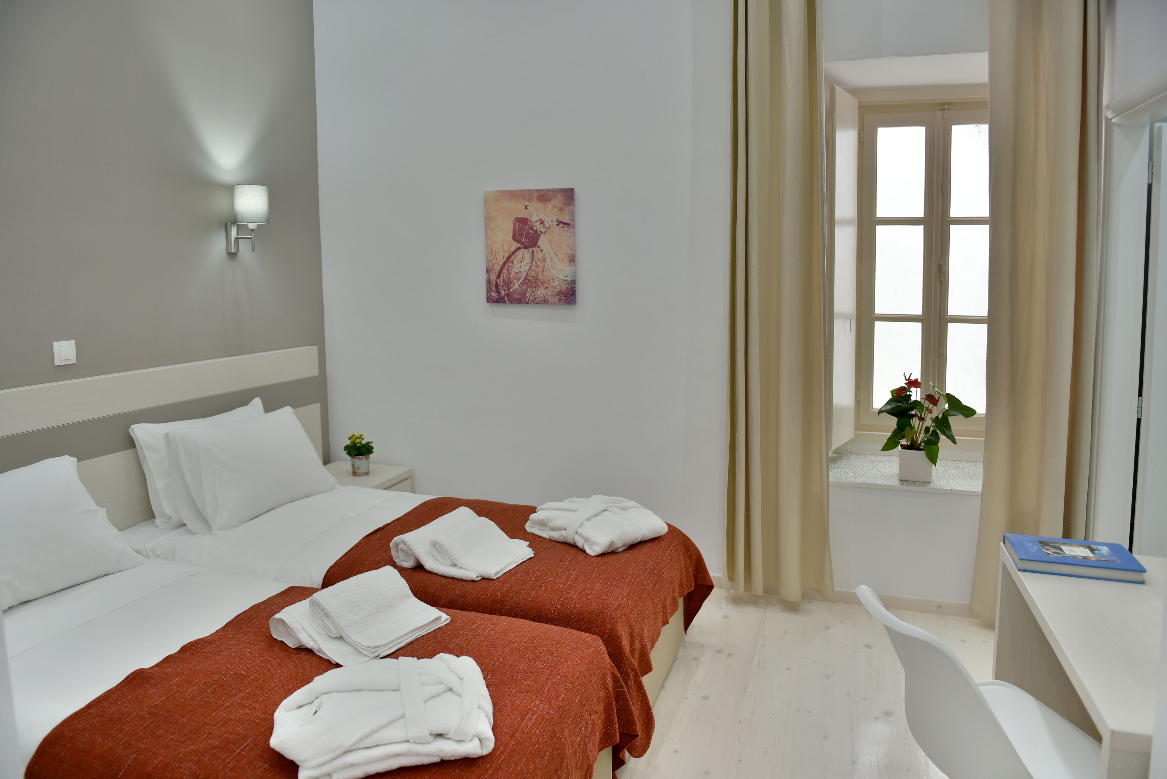 A hotel room with two beds, a desk, a chair, a window with curtains, and a wall-mounted light fixture.