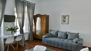 A modern hotel room with a grey sofa, a wooden wardrobe, a desk with chairs, a flat-screen TV, and a framed picture on the wall. - Douskos Port House (Hydra)