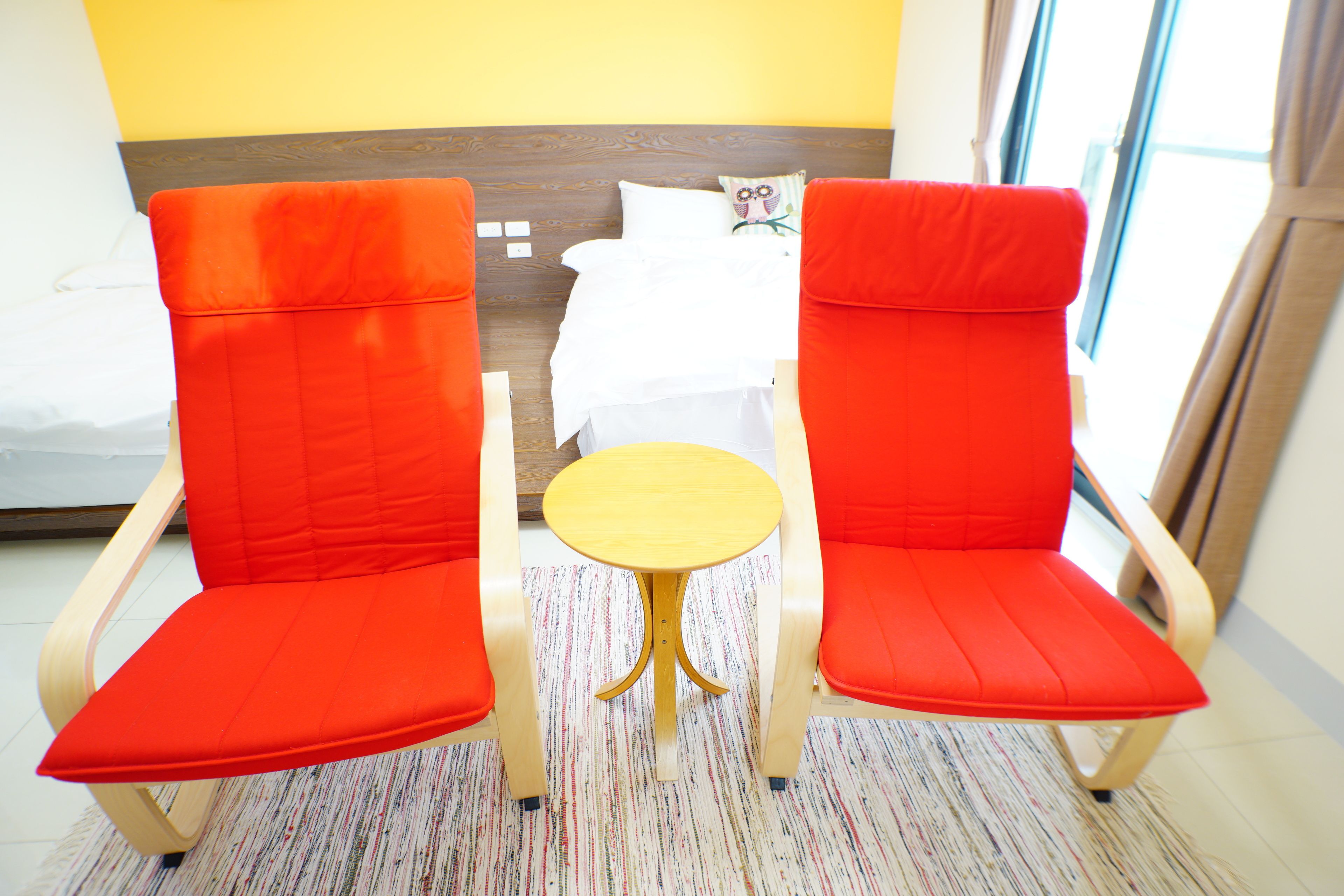 Una habitación de hotel con dos sillas rojas, una mesita redonda pequeña y una cama con ropa de cama blanca.