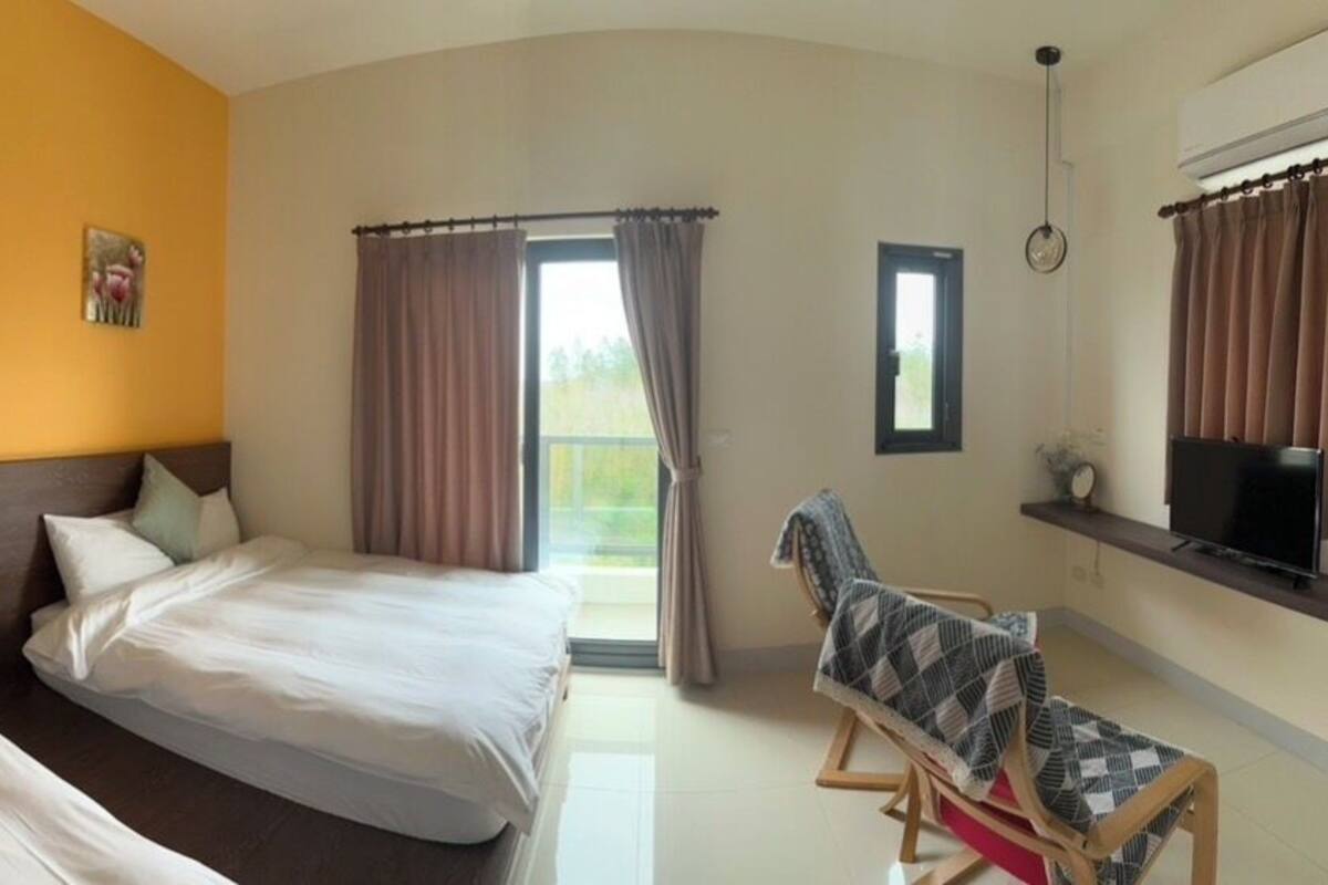 Ein Hotelzimmer mit zwei Betten, einem Fernseher, einem Sessel und einem Balkon mit Vorhängen.