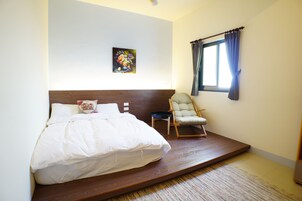 Un dormitorio con cama, una silla, una mesita, un cuadro y una ventana con cortinas.