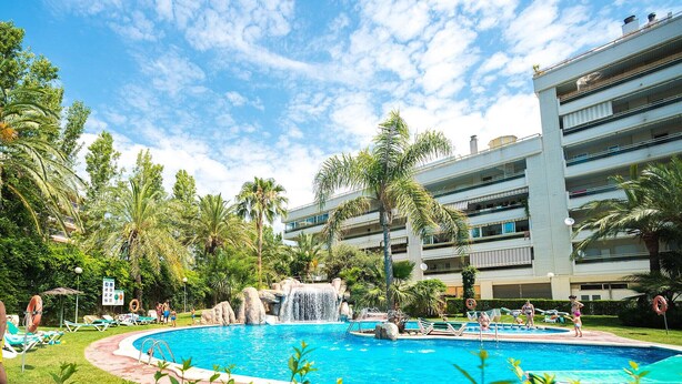 A pool area with a waterfall feature, surrounded by palm trees and lounge chairs. - PARAISOL 3 Family Complex (Salou)