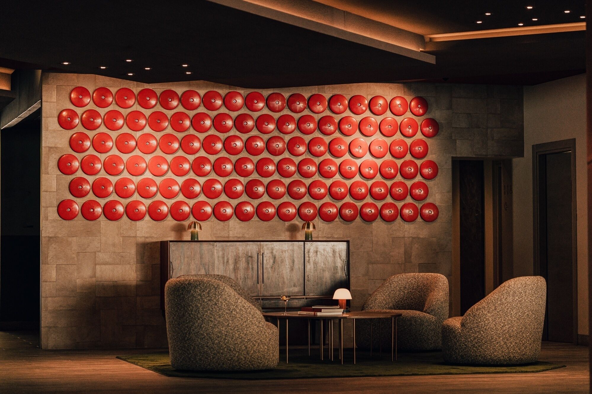Un interior moderno con una pared llena de objetos circulares rojos, un mueble de madera y dos sillas con textura.