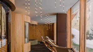 Een spa ruimte met houten wanden, een stenen vloer en een groot raam met uitzicht op een besneeuwd landschap. Er zijn verschillende houten ligbedden en een receptiegedeelte met een gebogen houten wand.