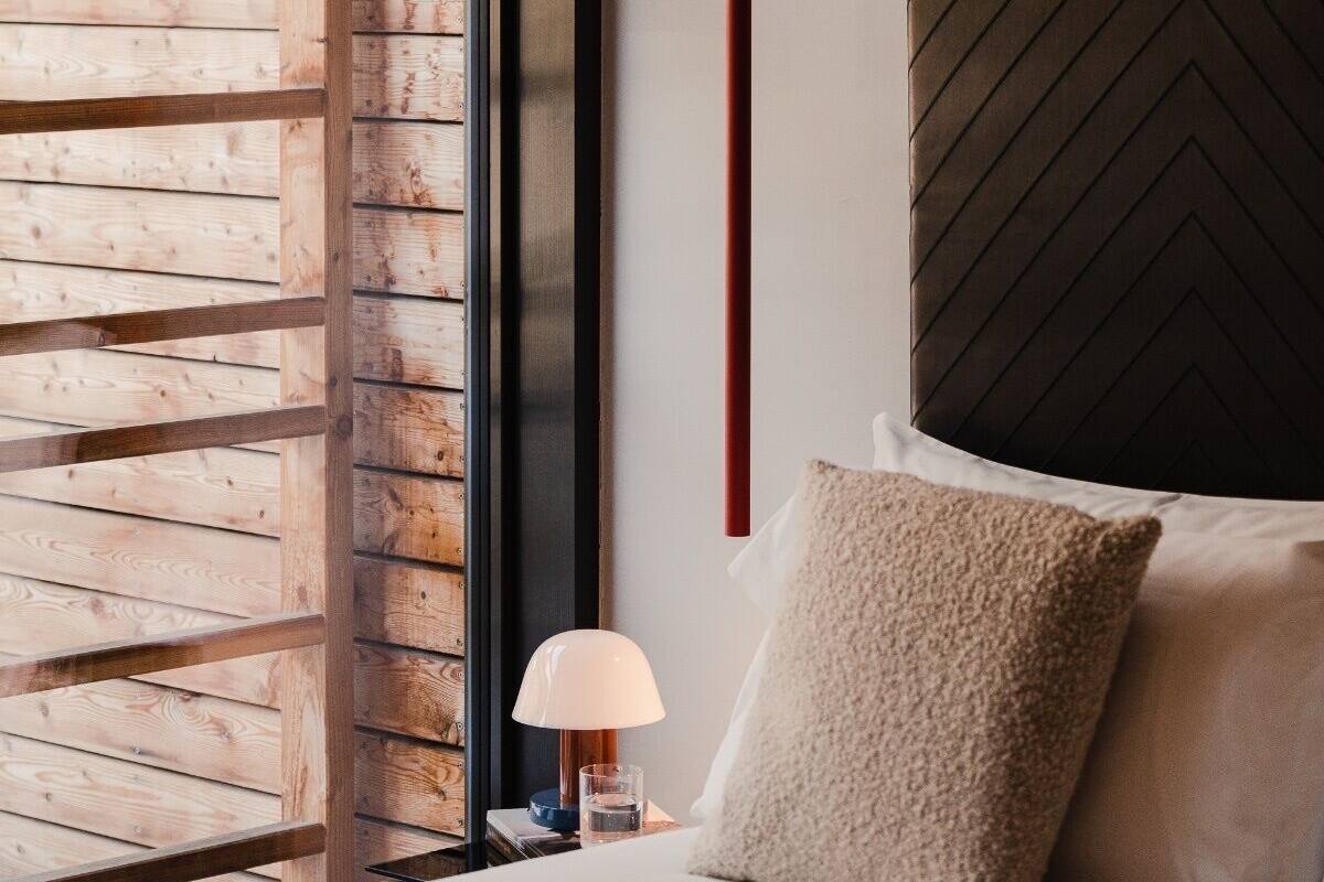 Un dormitorio con ventana de listones de madera, cama con cabecera de espiga y una lámpara de noche.