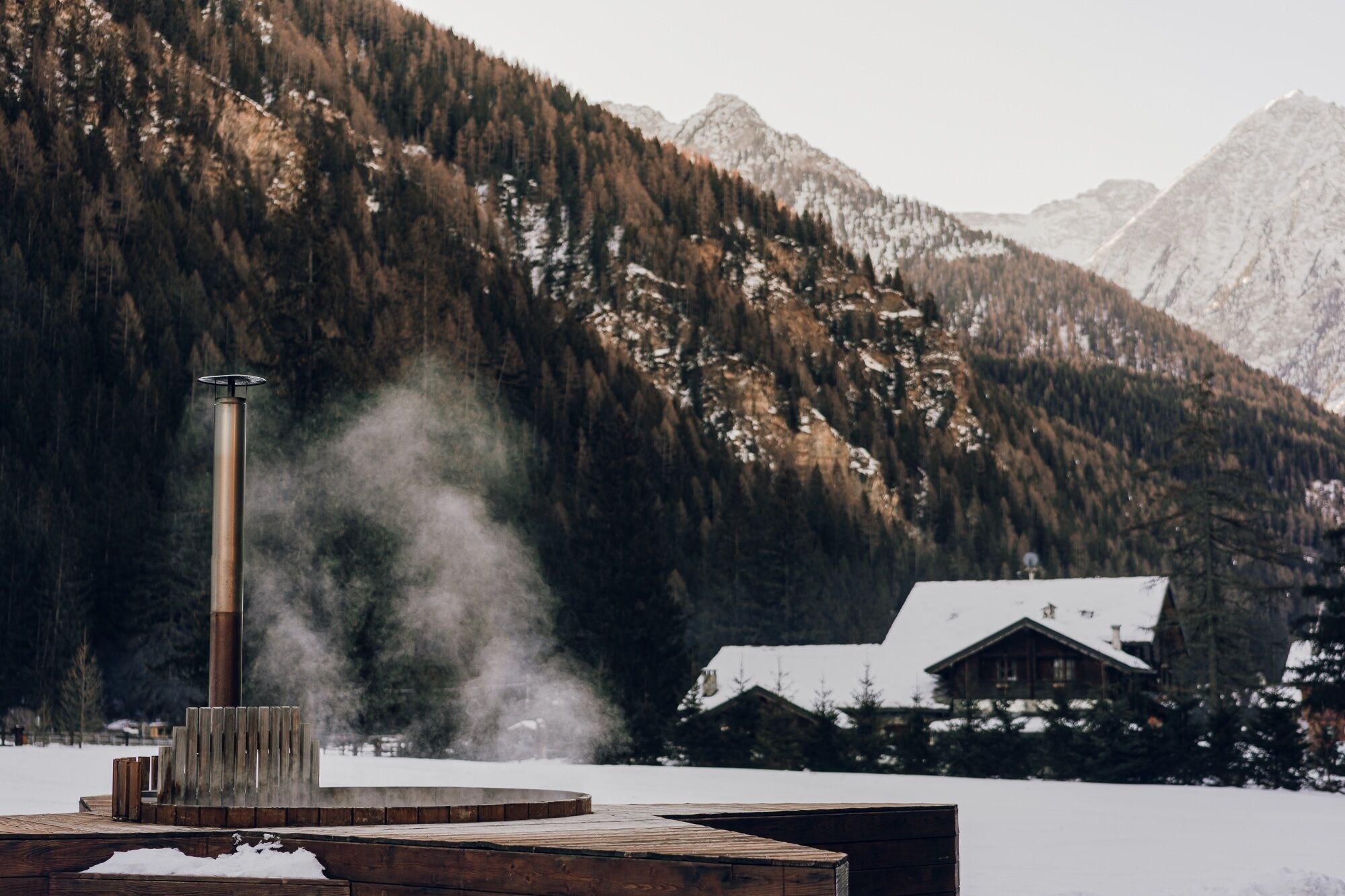Una sauna de madera con vapor ascendiendo, rodeada de nieve y un paisaje de montañas.