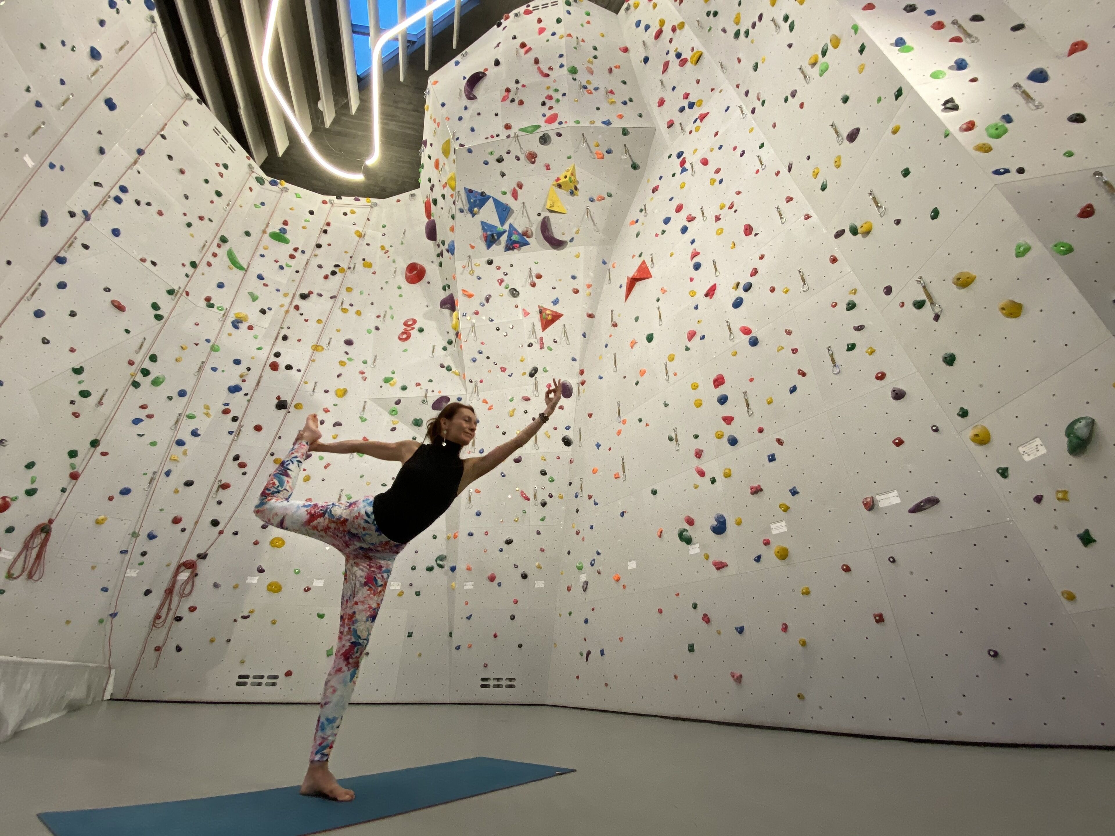 A person practicing yoga on a blue mat in a room with a climbing wall featuring various colored holds.