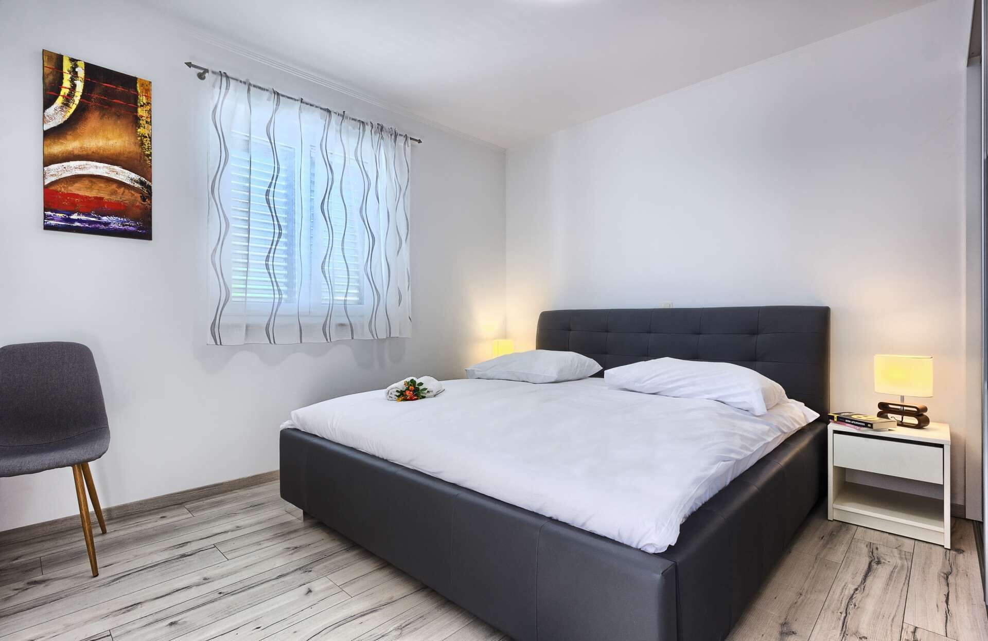 A modern bedroom with a bed, a chair, a bedside table with a lamp, and a window with curtains.