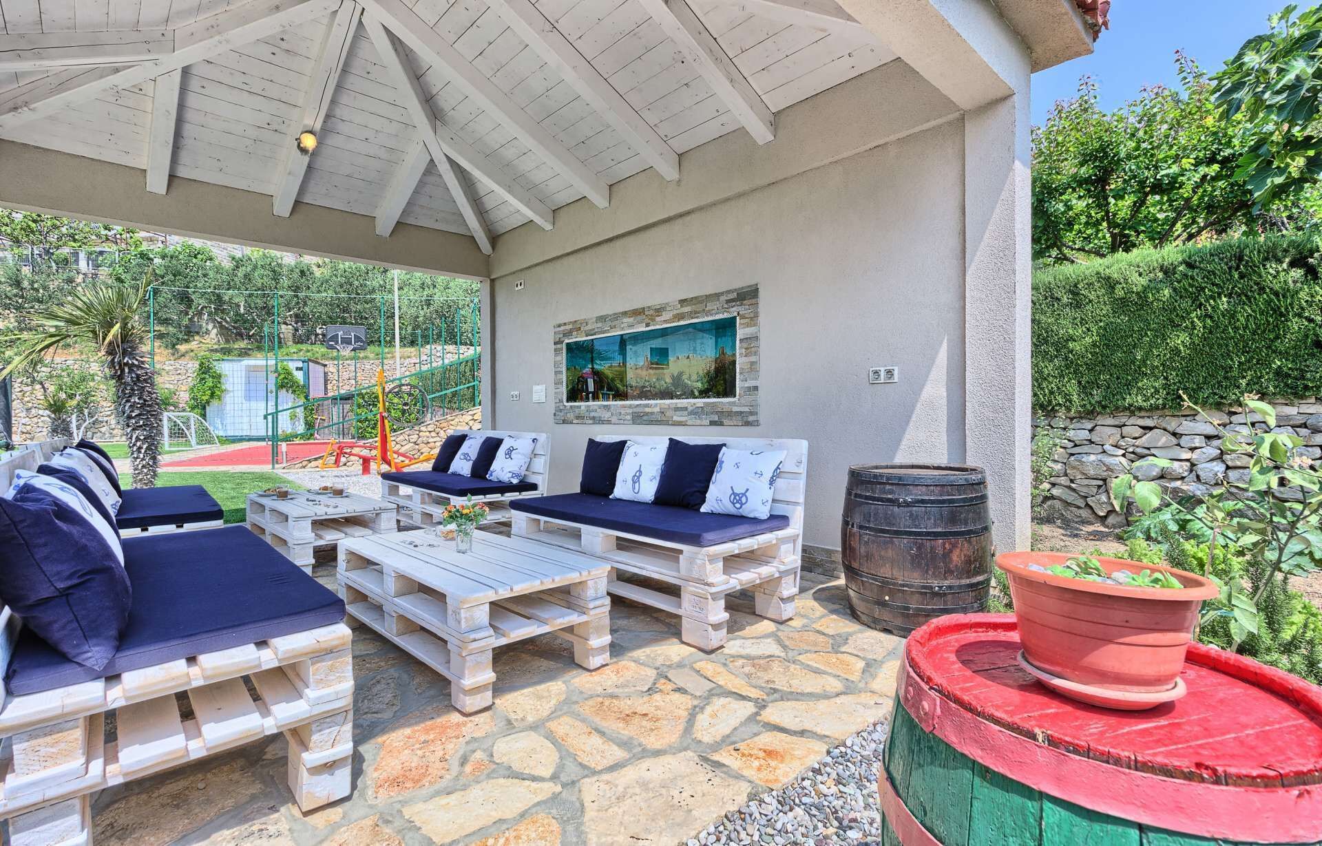 An outdoor patio with wooden furniture, a stone floor, a painting on the wall, and a barrel.