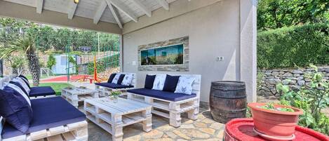 An outdoor patio with wooden furniture, a stone floor, a painting on the wall, and a barrel.