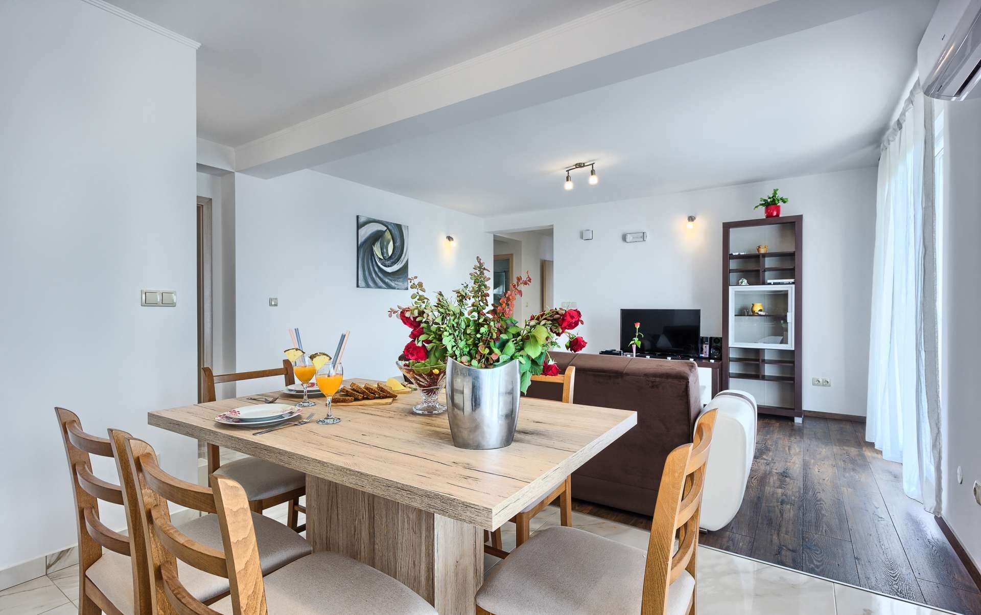 Un coin repas avec une table en bois, des chaises et un vase de fleurs. On aperçoit un téléviseur et une bibliothèque à l’arrière-plan.