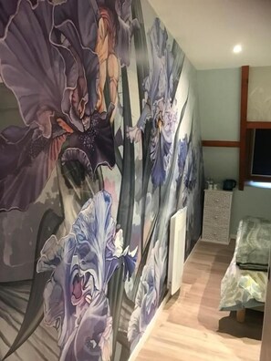 A room with a large floral mural on the wall, a bed with a patterned bedspread, and a window with a view of the outside. - Chambre d'hôtes Mon Taj Mahal (Gueron)