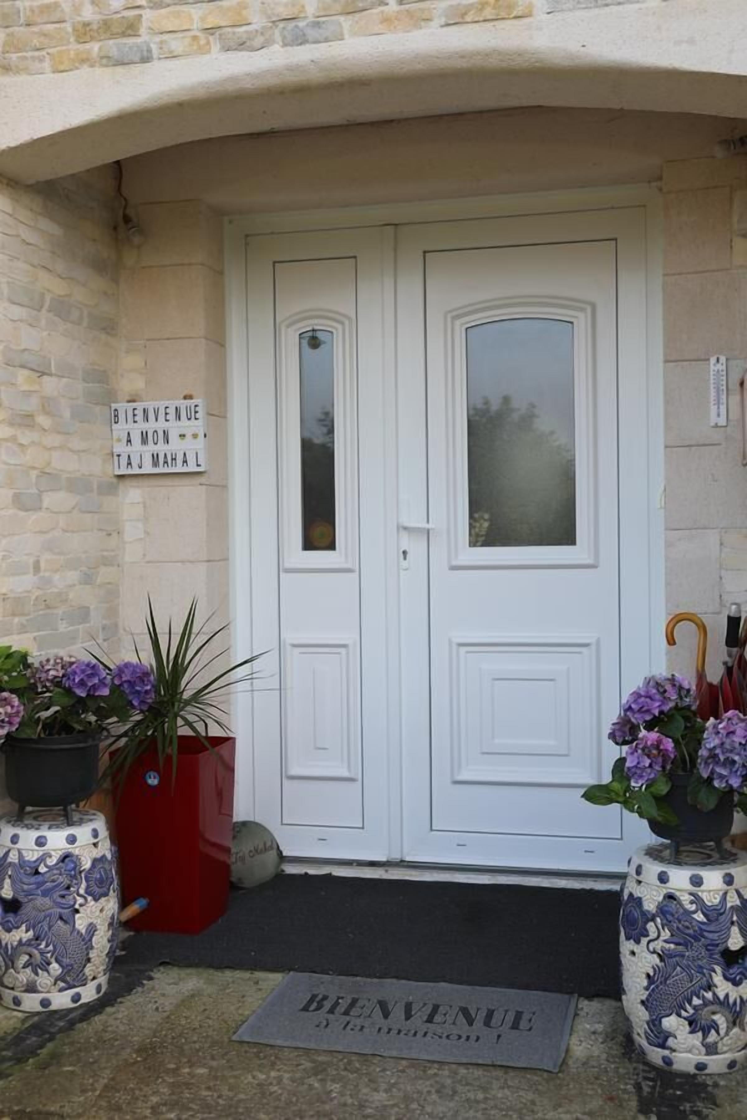 Una puerta doble blanca con un panel de vidrio satinado, flanqueada por dos macetas con flores moradas. Un letrero de bienvenida está colgado en la pared a la izquierda.