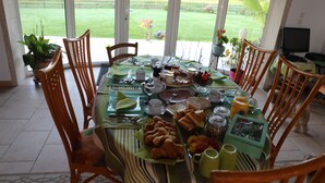 A dining table set for breakfast with various food items and drinks, surrounded by wooden chairs, with a view of a garden through large windows. - Chambre d'hôtes Mon Taj Mahal (Gueron)