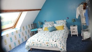 A bedroom with a sloped ceiling, a bed with patterned bedding, a nightstand, a chair, and a shelf with a stuffed toy. - Chambre d'hôtes Mon Taj Mahal (Gueron)