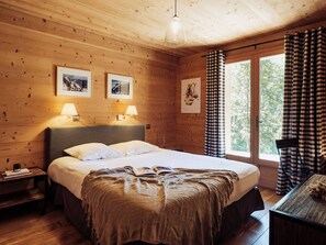 A wooden cabin bedroom with a bed, two bedside lamps, framed pictures on the wall, and a window with checkered curtains.