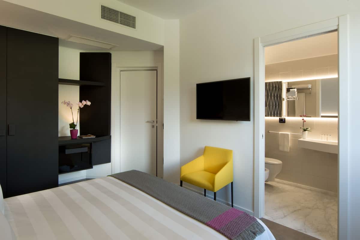 A hotel room with a bed, a TV mounted on the wall, a small yellow chair, and a bathroom visible through an open door.