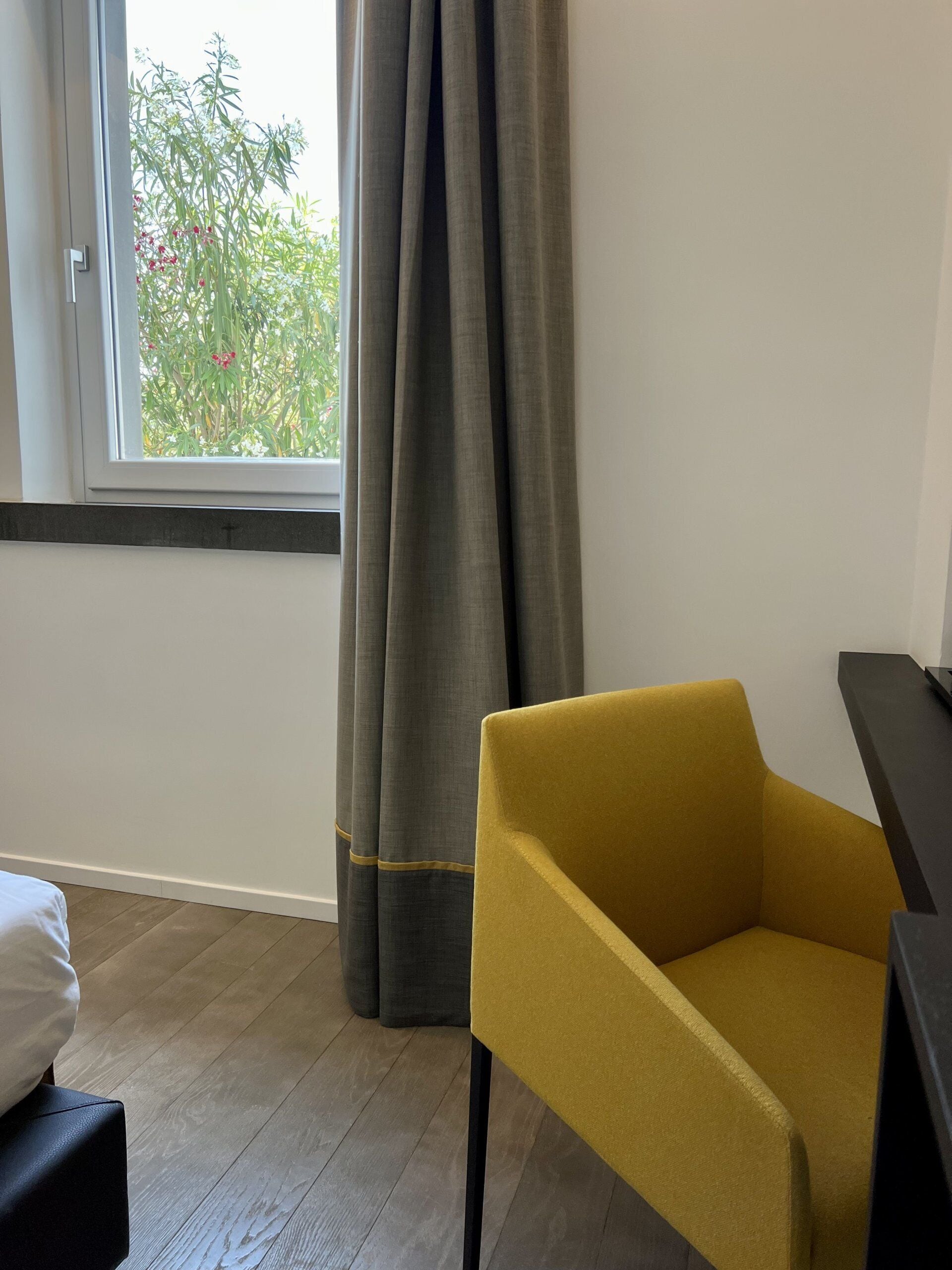 A room with a window, grey curtains, a yellow chair, a black table, and a white pillow on a black ottoman.
