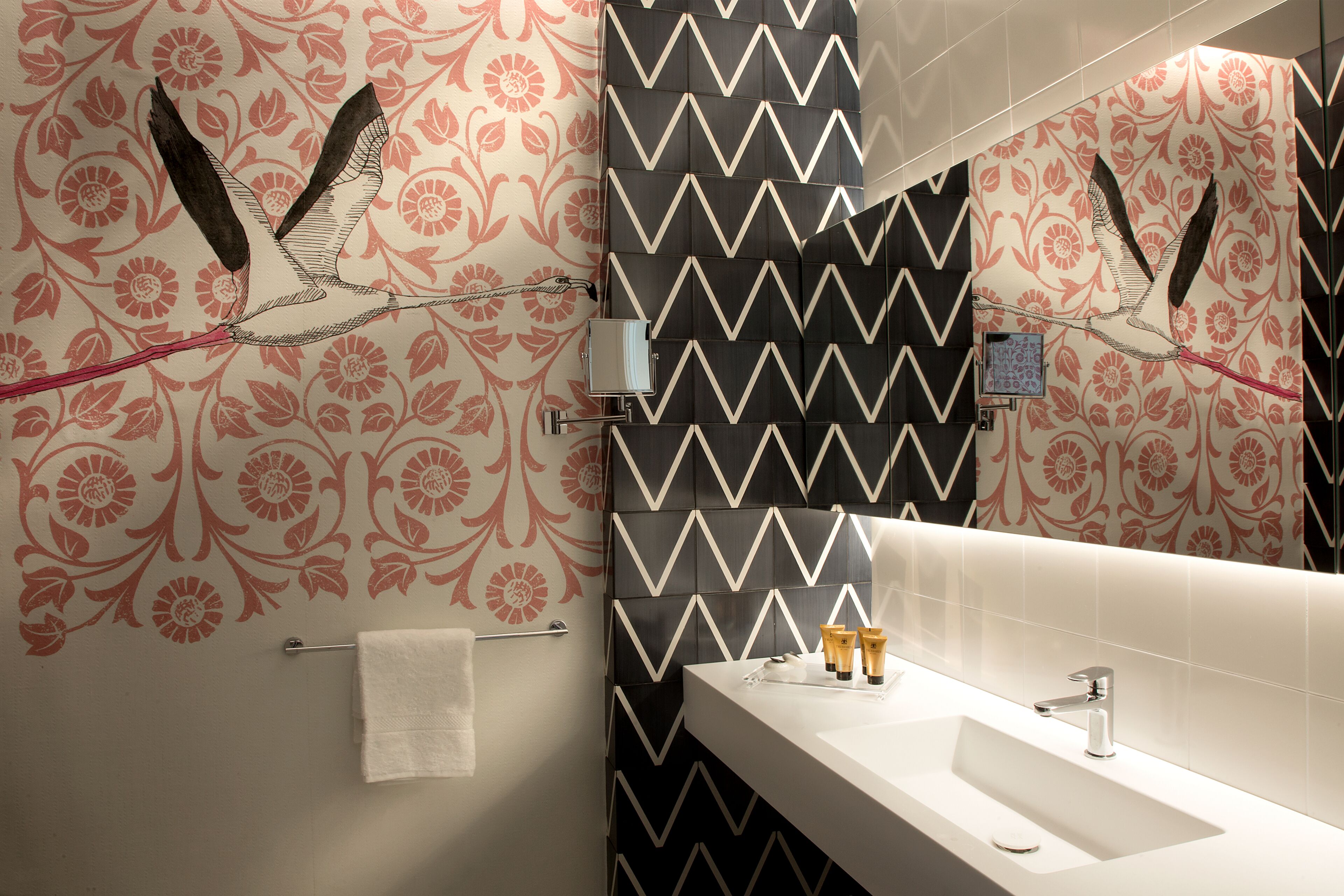 Une salle de bain avec un lavabo blanc, un miroir et un porte-serviettes. Les murs sont ornés d’un motif floral et d’un motif en zigzag noir et blanc.