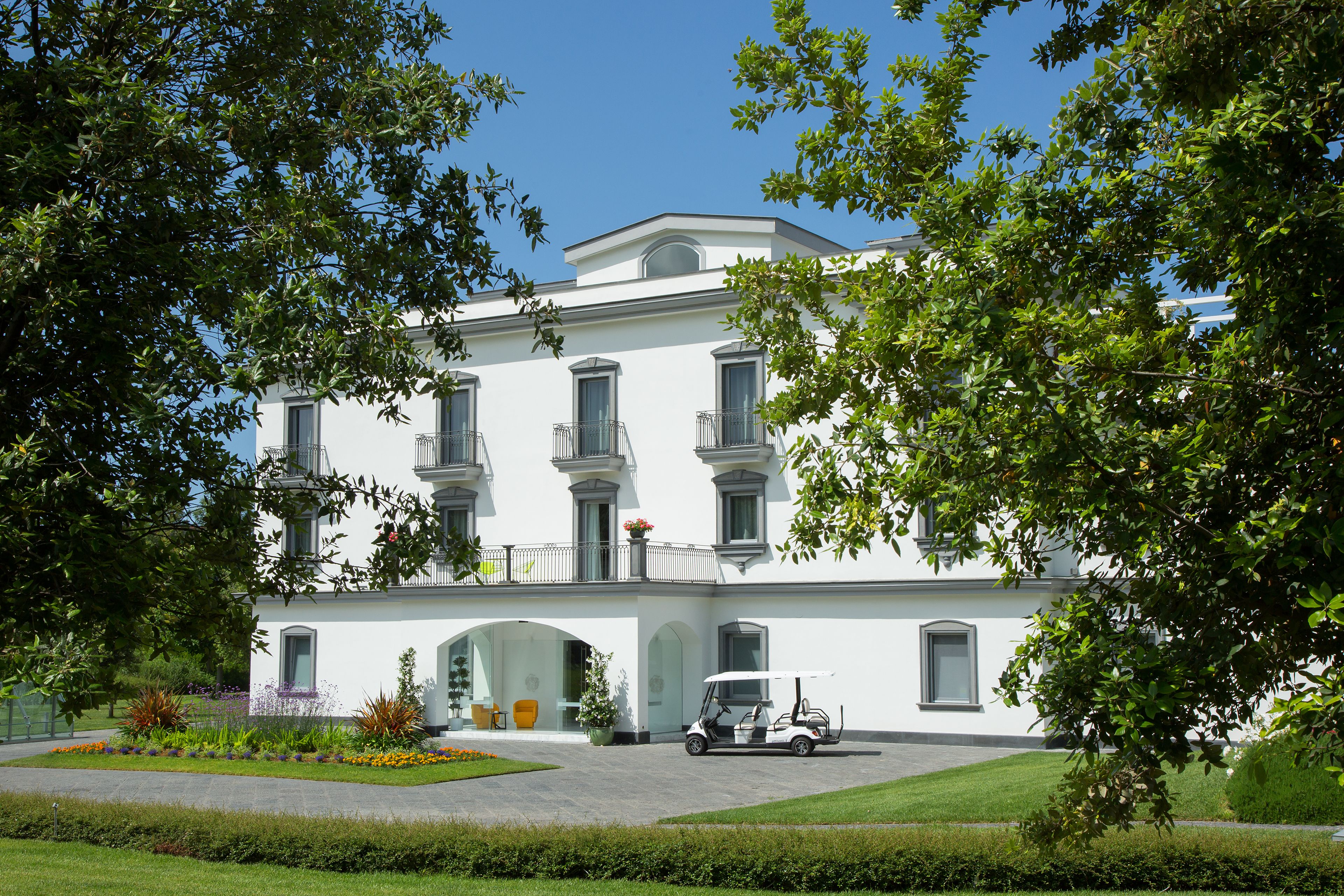 A white building with a balcony, surrounded by greenery and a golf cart in front.