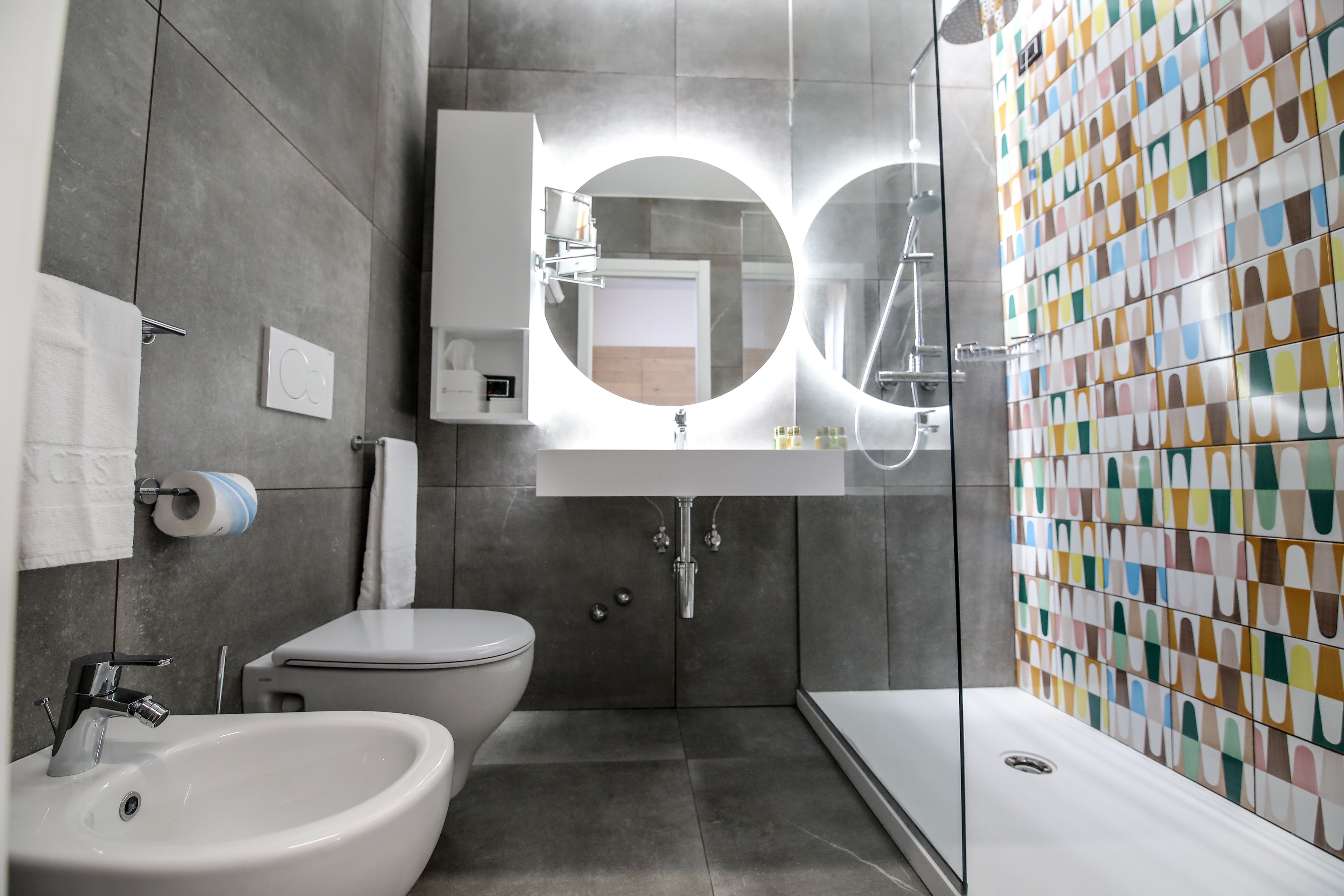 A modern bathroom with a white sink, toilet, and shower. It features a large mirror with a circular light above it and a colorful tile wall.