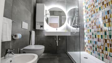 A modern bathroom with a white sink, toilet, and shower. It features a large mirror with a circular light above it and a colorful tile wall.