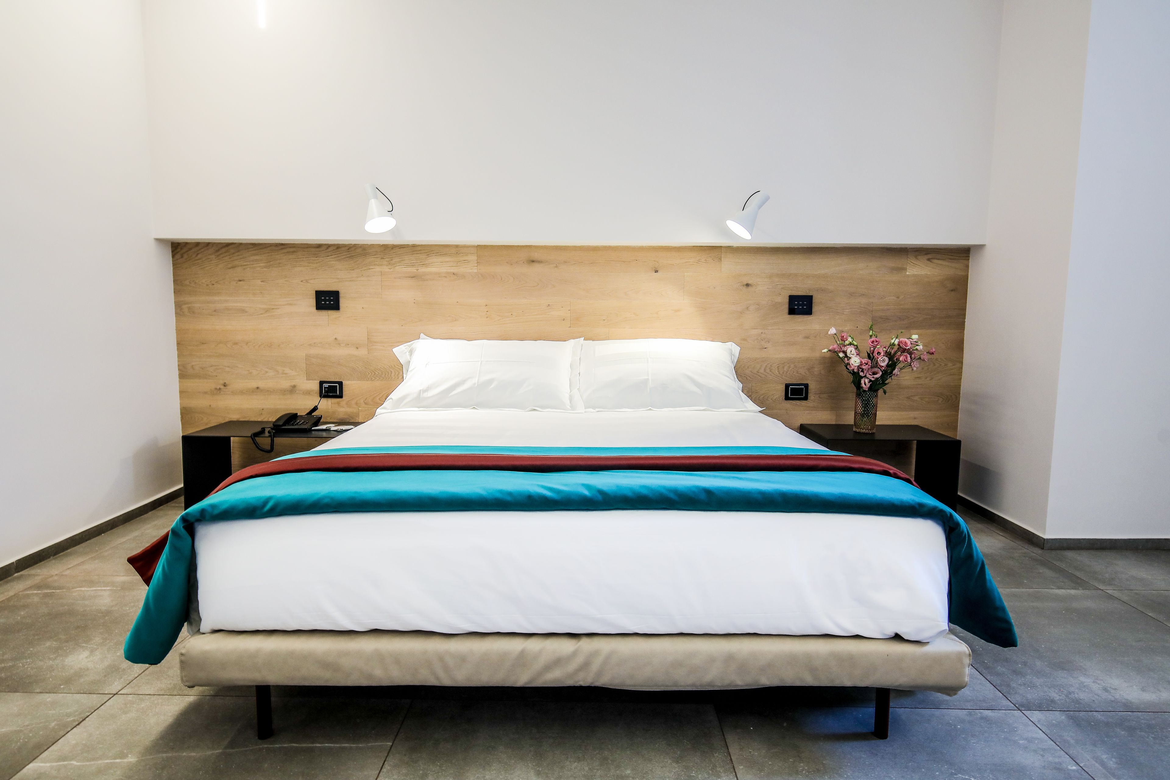 Une chambre d’hôtel moderne avec une tête de lit en bois, un lit bien fait avec une literie blanche et turquoise, et une petite table de chevet sur laquelle se trouvent un téléphone et un vase de fleurs.