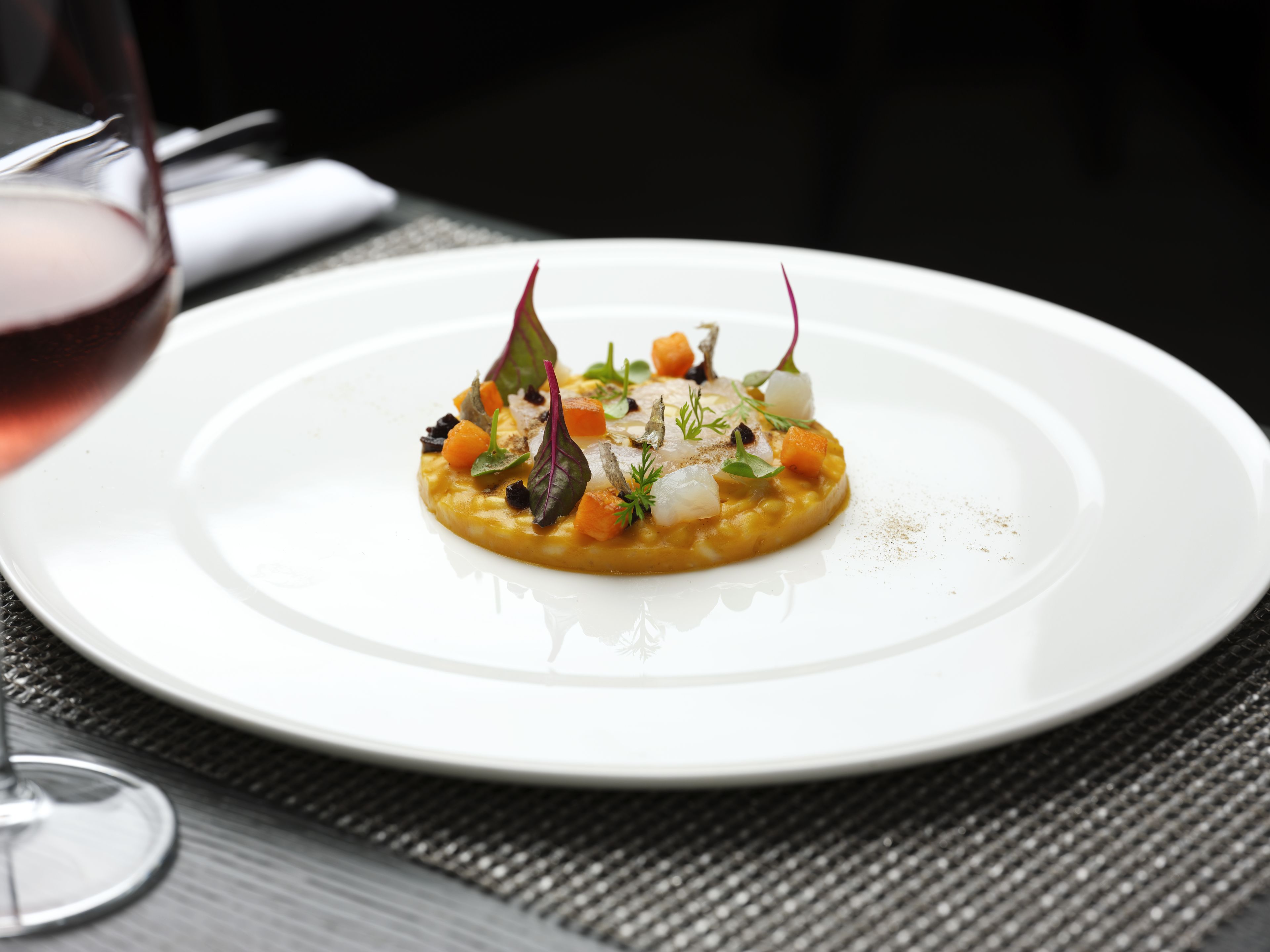 A white plate with a gourmet dish, a glass of red wine, and a napkin on a table.