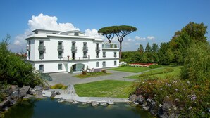 A large white building with multiple windows and a central entrance, surrounded by a well-maintained garden and a pond. - Il San Cristoforo (Ercolano)
