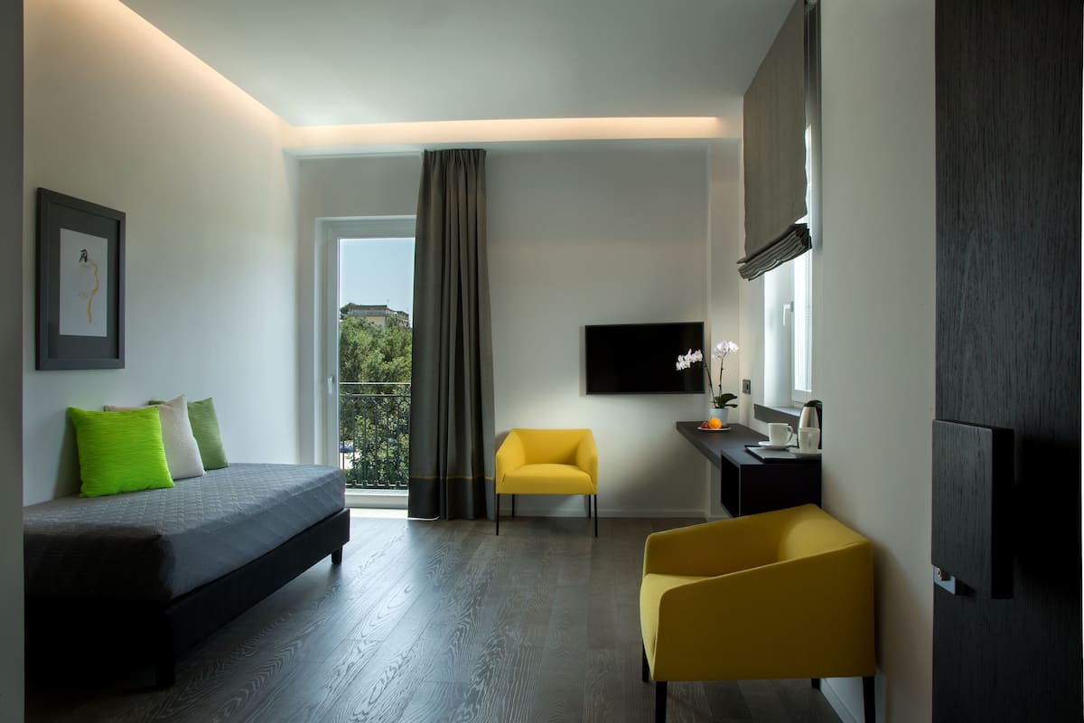 A modern hotel room with a bed, a yellow armchair, a desk with a flat-screen TV, and a large window with curtains.
