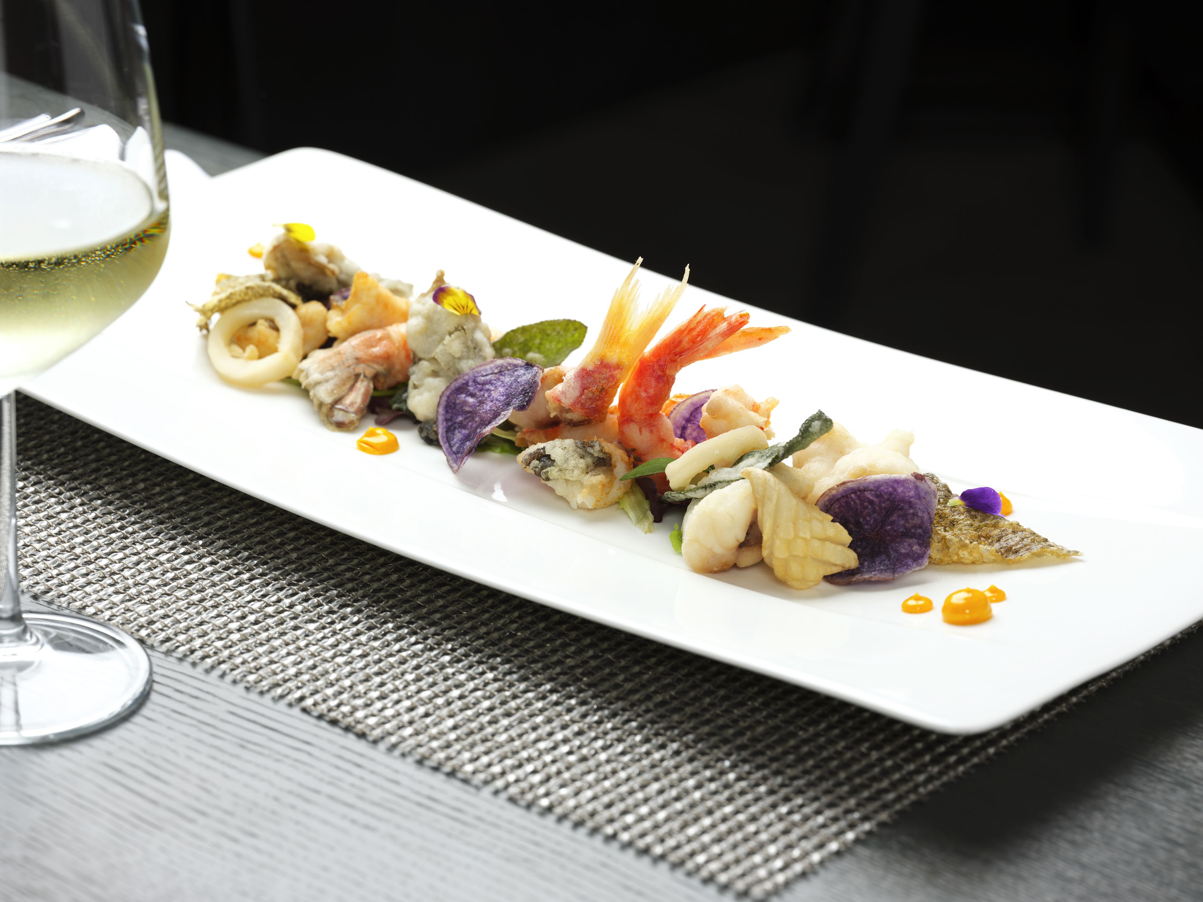 Un plato blanco rectangular con un plato de mariscos, una copa de vino blanco y un mantel tejido.