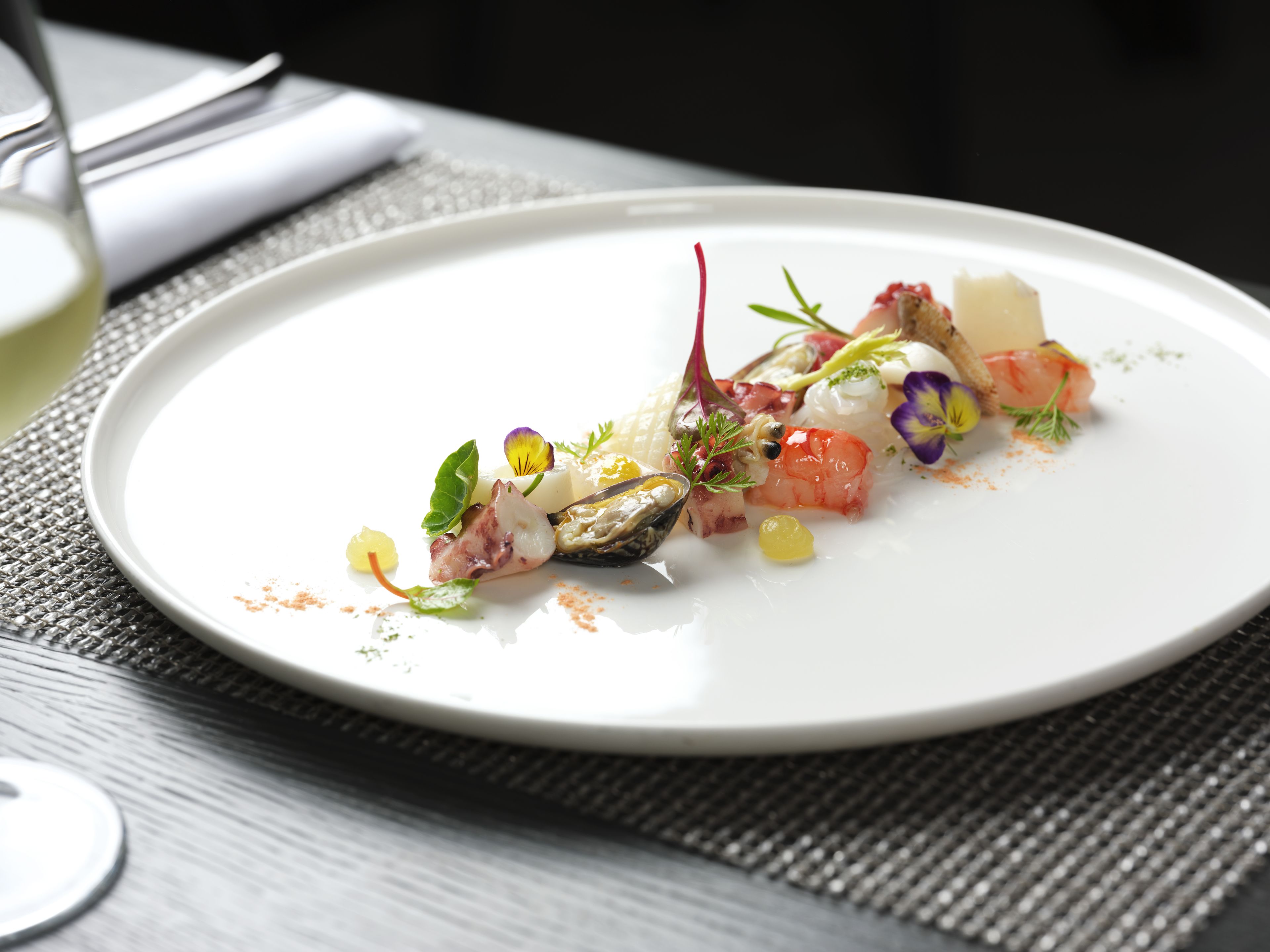 Une assiette blanche contenant un plat de fruits de mer, agrémenté d’herbes aromatiques et de fleurs comestibles, posée sur une nappe à motifs.