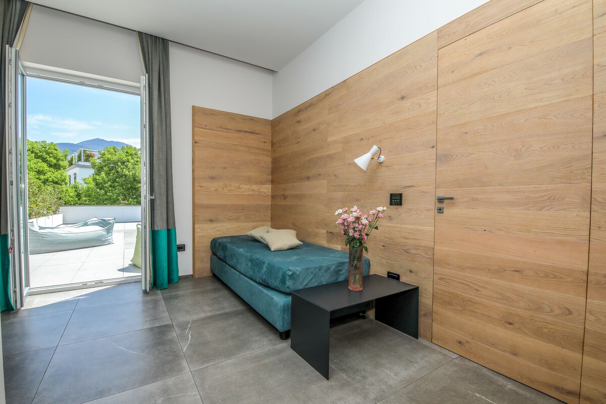 Un dormitorio moderno con sofá cama, una mesita de noche con un florero y vista a un paisaje verde a través de una puerta corredera.