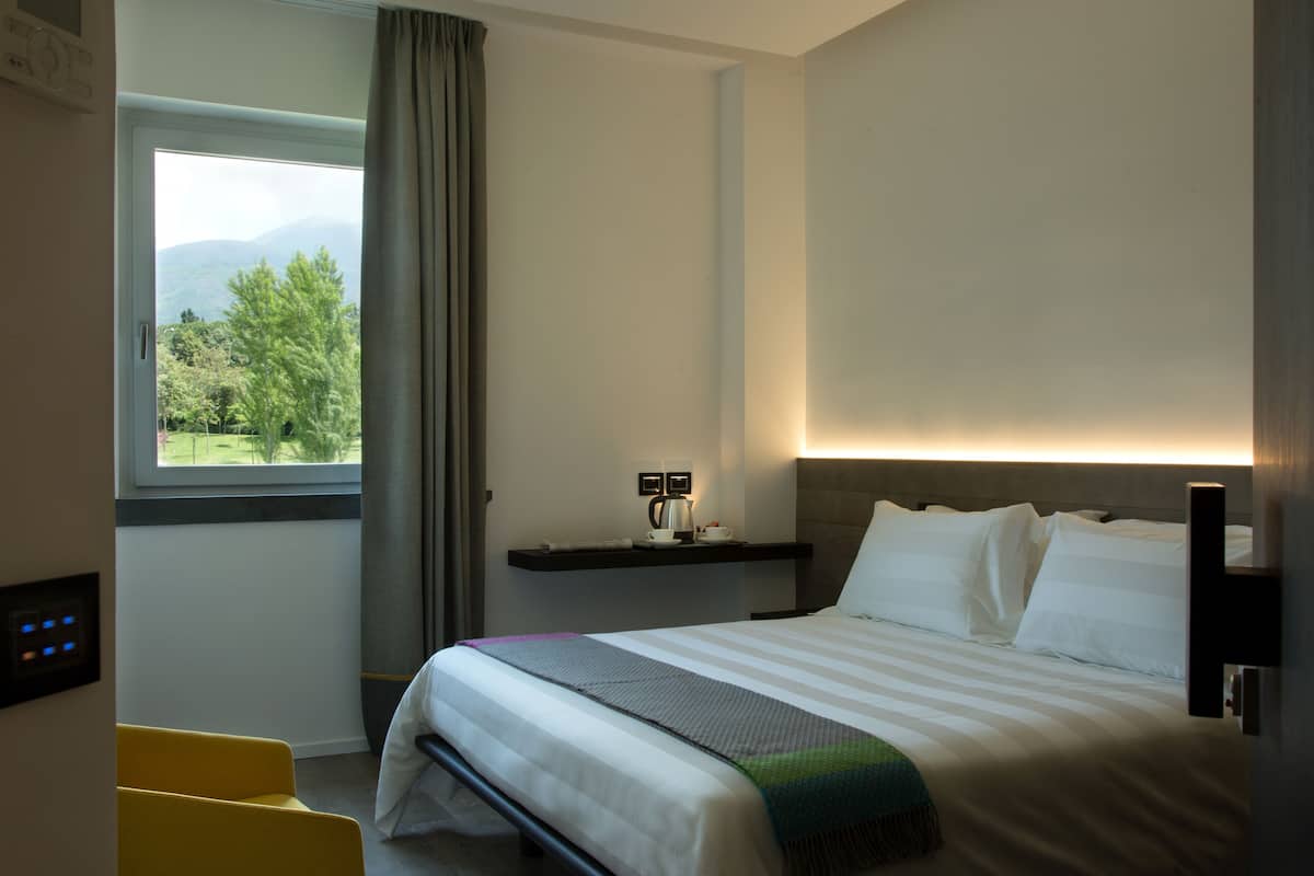 A hotel room with a bed, a desk, a chair, a window with a view of trees and mountains, and a shelf with a kettle and cups.