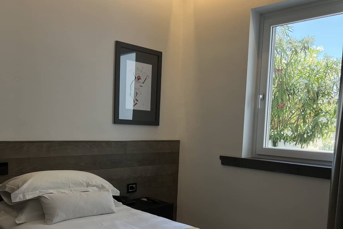 A neatly made bed with white linens and a beige throw, a bedside table with a lamp, and a window with a view of greenery.