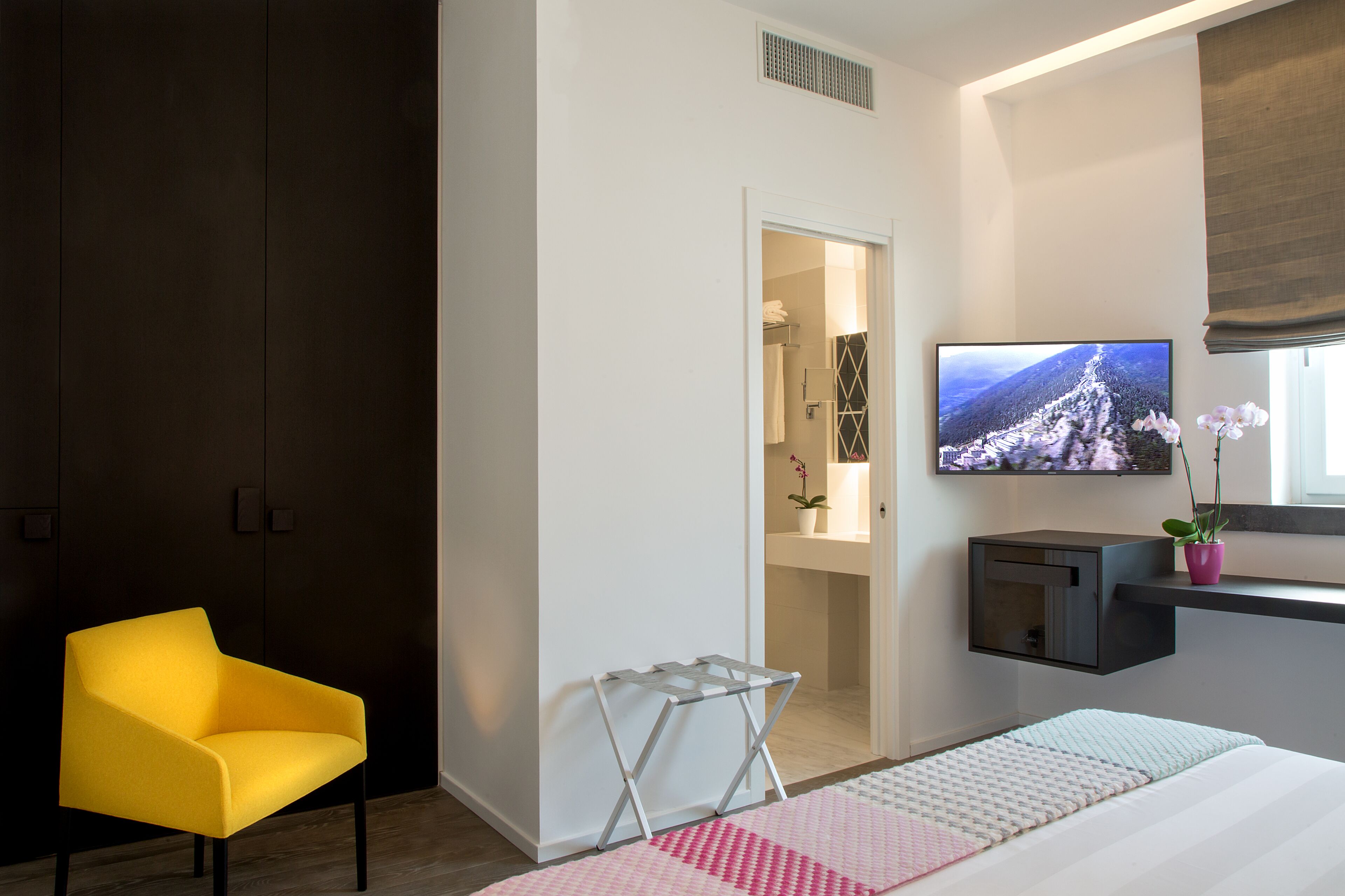 Una habitación de hotel moderna con una cama, un sillón amarillo, un televisor y una mesita.