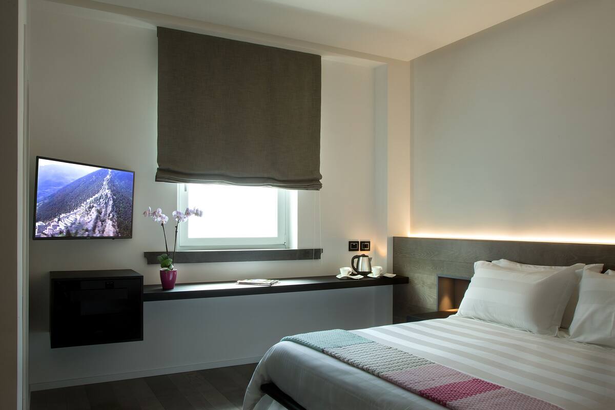 A modern hotel room with a bed, a TV mounted on the wall, a window with a view, a bedside table with a phone and a cup, and a small desk with a computer.