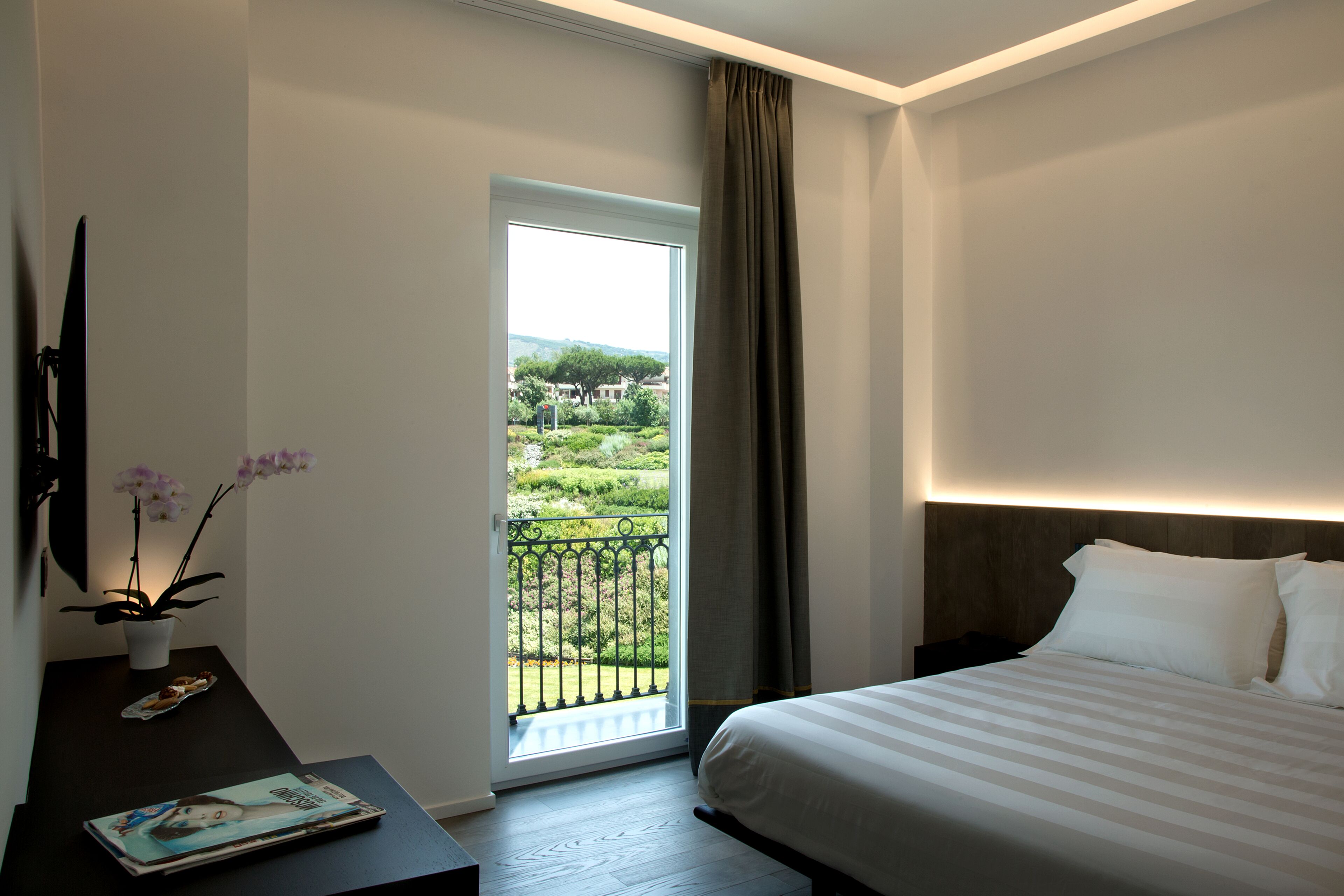 Una camera d'albergo con un letto, una scrivania con una rivista, una pianta in vaso e vista su un giardino tramite una porta aperta.