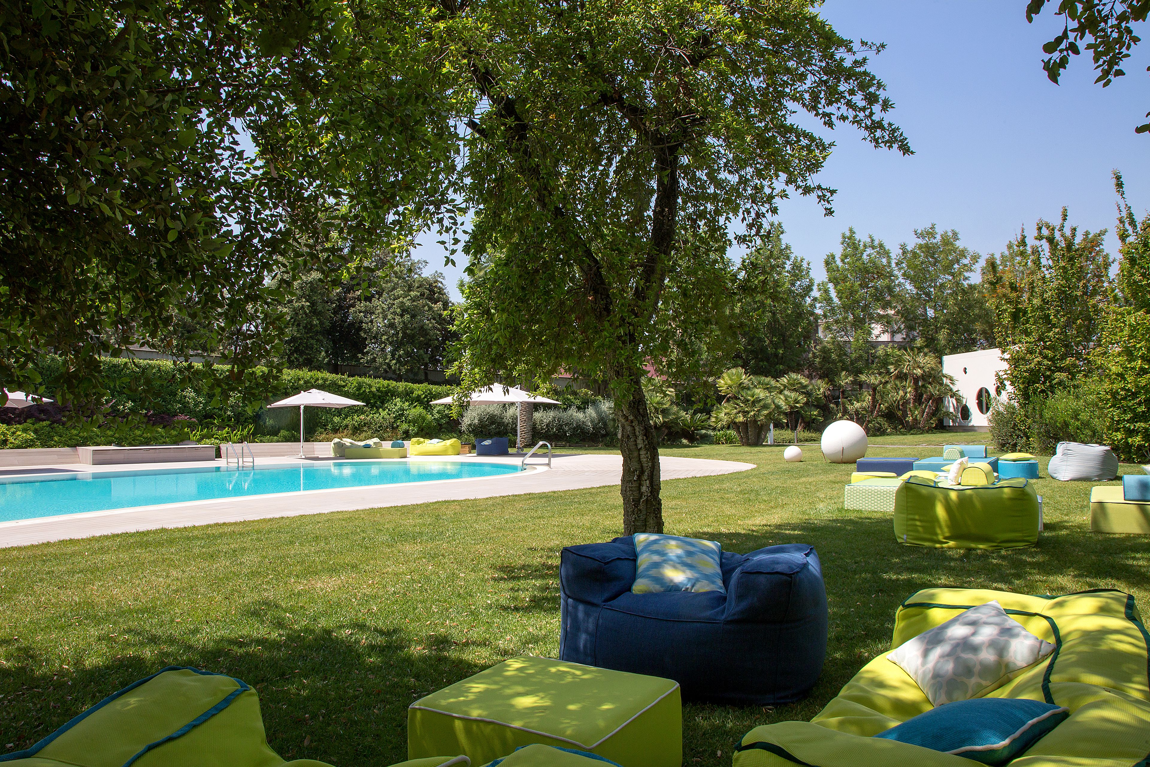 Un área junto a la piscina con piscina, sillas de descanso verdes y azules, y un árbol que da sombra.
