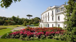 A large white building with multiple windows and balconies, surrounded by lush greenery and vibrant flower beds. - Il San Cristoforo (Ercolano)