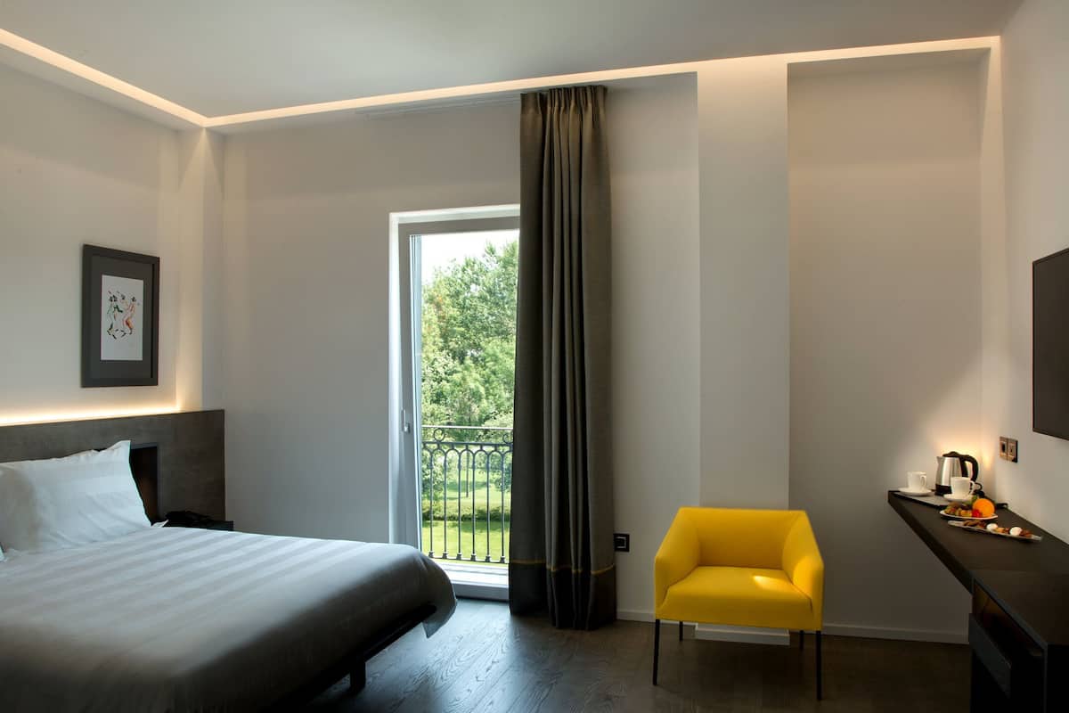 A hotel room with a bed, a desk with breakfast, a chair, and a balcony with curtains.