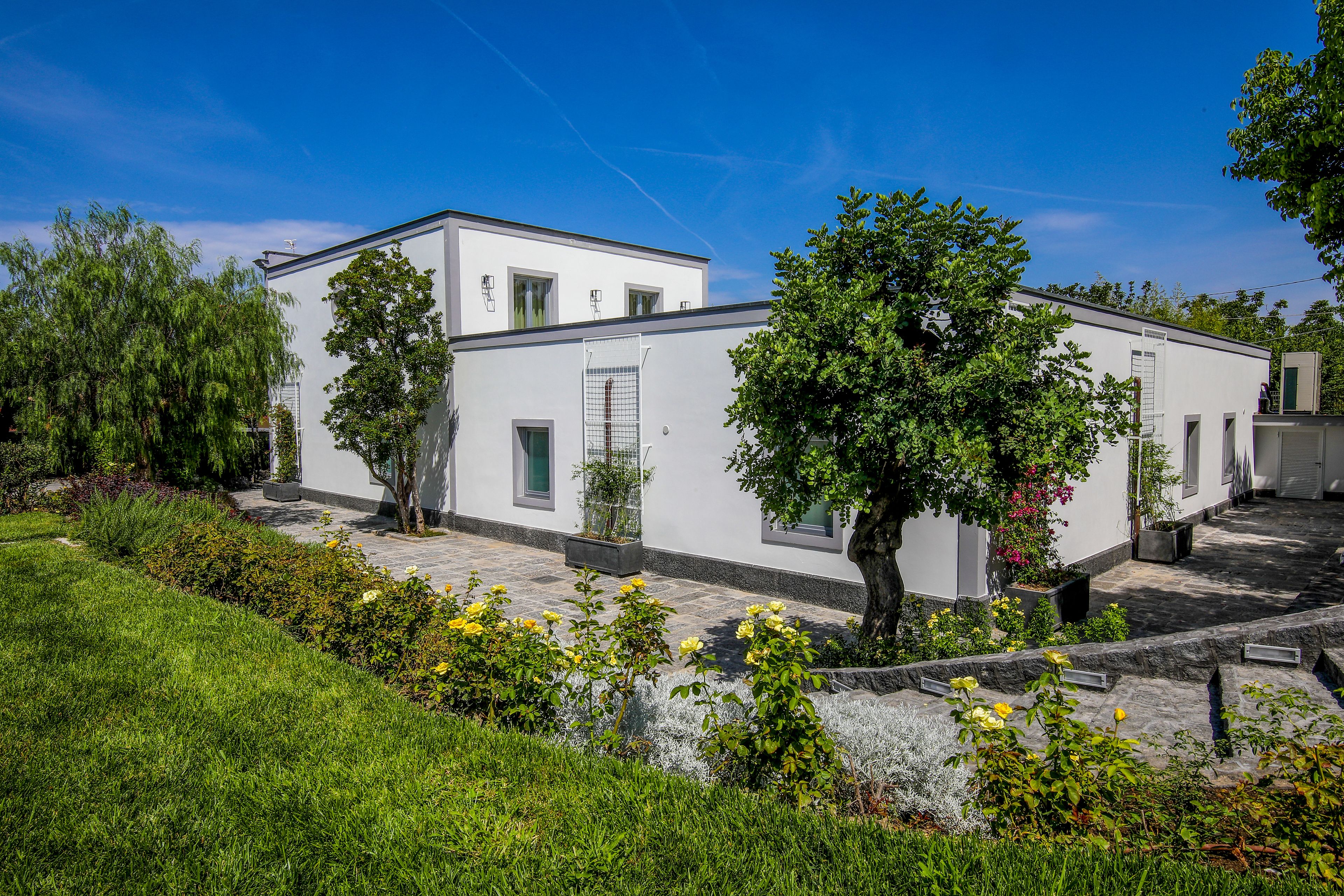 Un bâtiment moderne aux murs blancs, avec un chemin en pierre, entouré d’une végétation luxuriante et de plantes en fleurs.