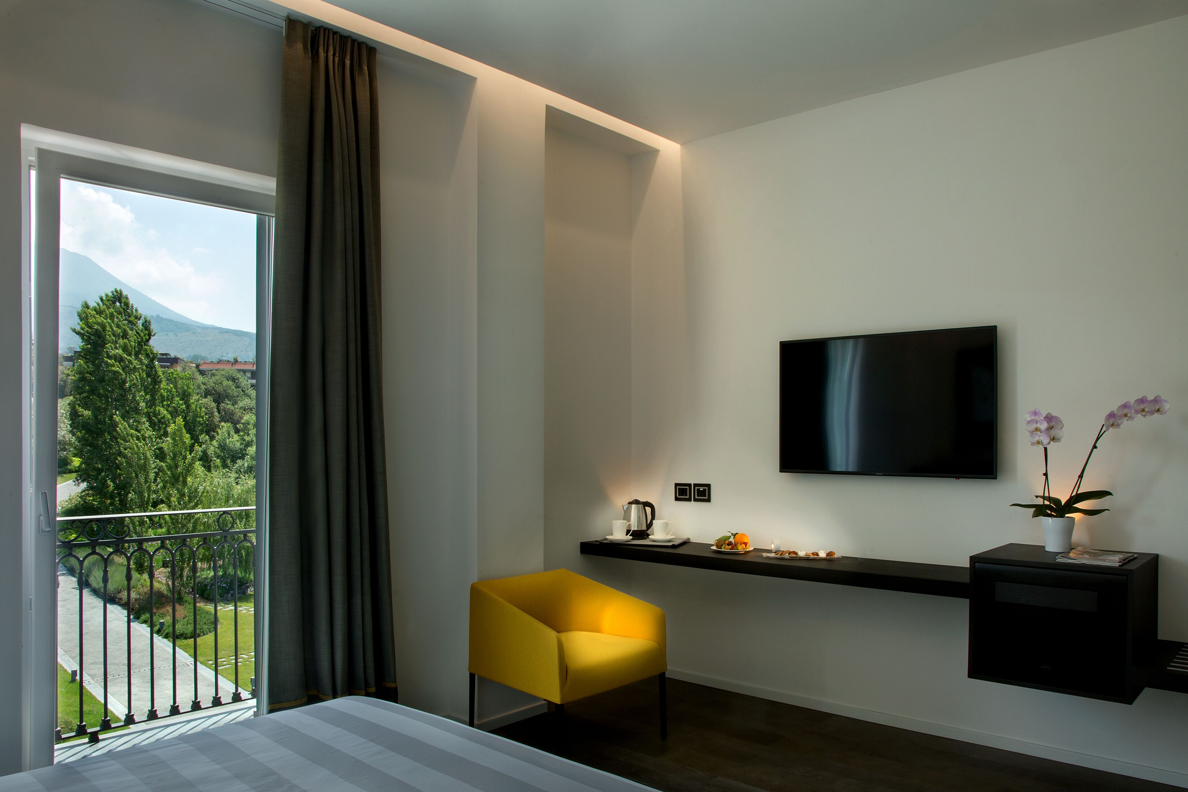 A hotel room with a bed, a yellow chair, a TV, a small table with a plant, and a view of greenery and mountains.