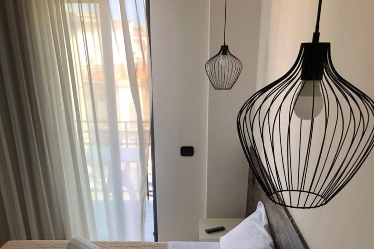 A hotel room with two beds, a large window with sheer curtains, a hanging black pendant light, and a small bedside table with a remote control.