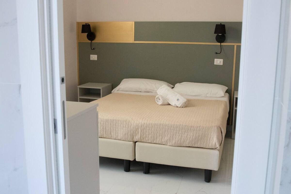 A modern hotel room with a large bed, two bedside tables, and a headboard with wall-mounted lamps.