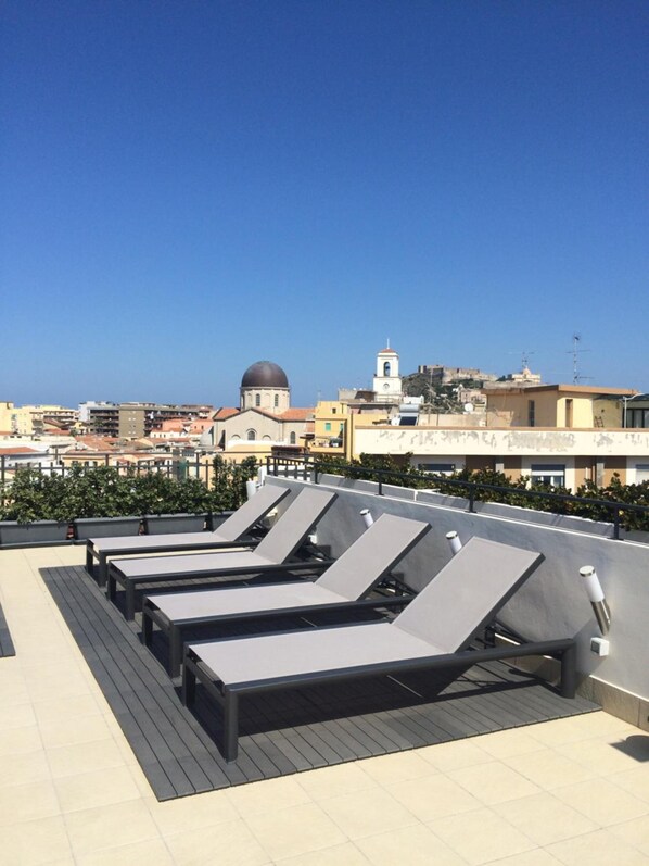 A rooftop terrace with sun loungers, a clear blue sky, and a cityscape with buildings and a dome in the background. - Milazzo INN AiMori (Milazzo)